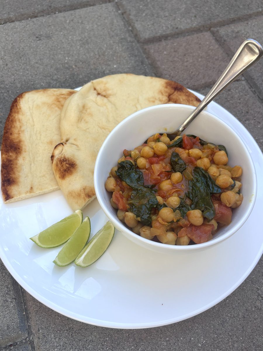 Chickpea, spinach and tomato curry in a white bowl with a spoon, plated with two pieces of naan and lime wedges on a white plate r