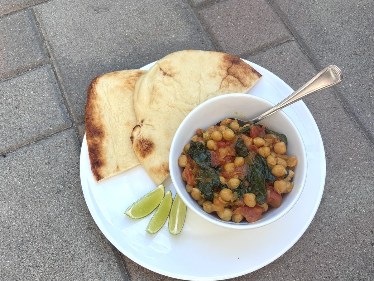 Overhead of the same chickpea and spinach curry in a white bowl with spoon, naan and lime wedges on a white plate set on outdoor p