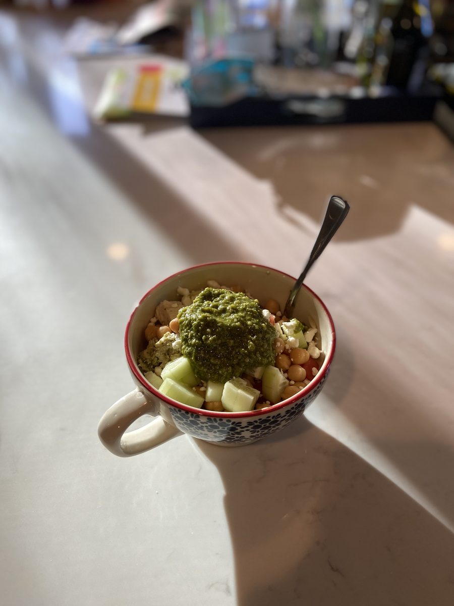 A small handled ceramic bowl of chickpeas with diced cucumber and crumbled feta topped with a spoonful of green pesto and a spoon,