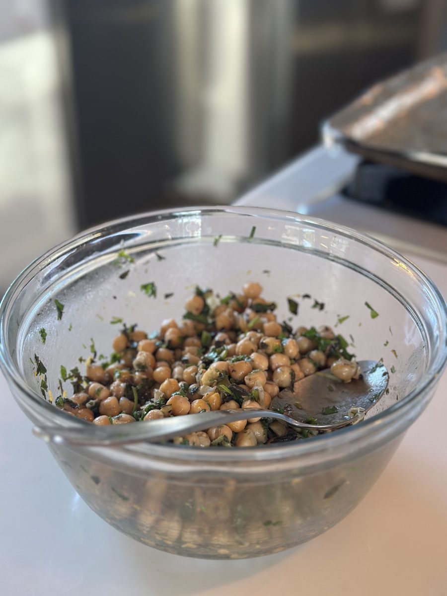 Chickpeas tossed with chopped fresh herbs and a spoon in a clear glass mixing bowl on a white counter, with a dark stove and a met