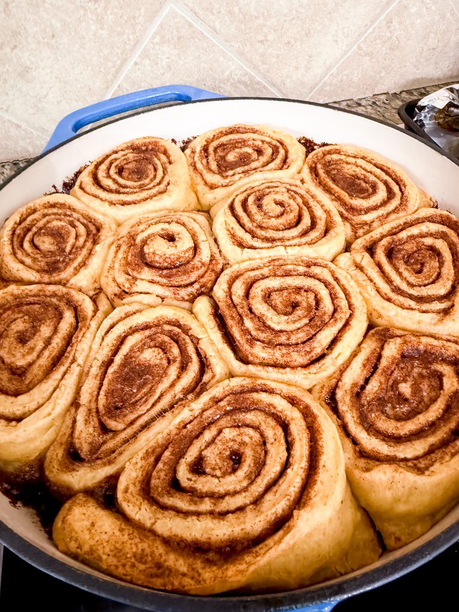 Golden baked cinnamon rolls, unfrosted, filling a blue enameled dutch oven photographed from above against a tiled backdrop.