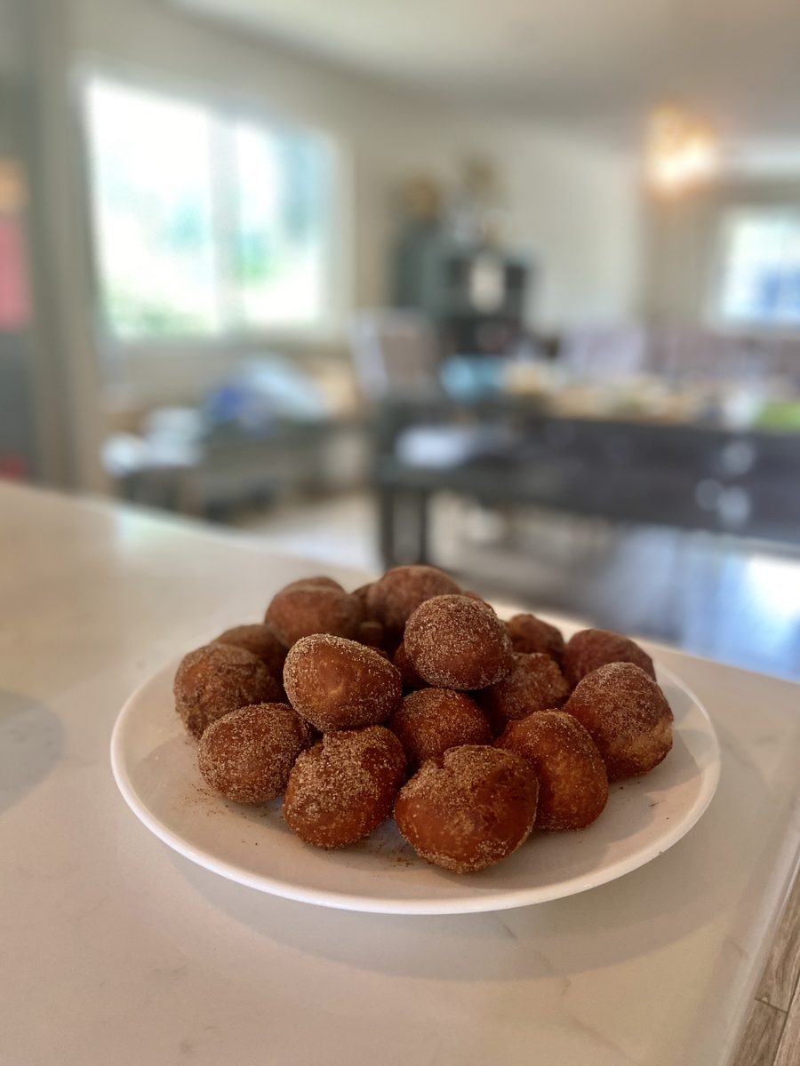 A white plate stacked with round cinnamon-sugar coated donut holes on a marble kitchen island, with a blurred open-plan living roo