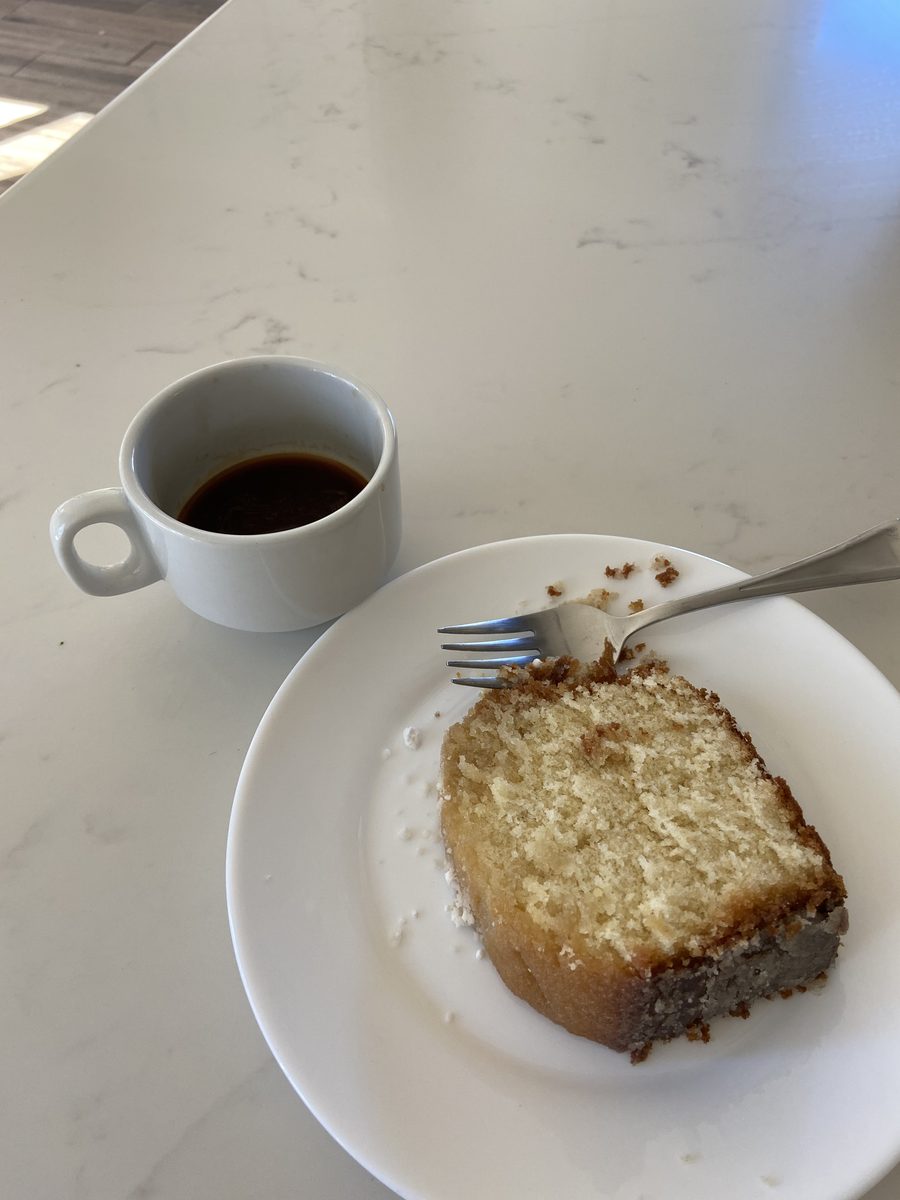 A slice of plain pale cake on a white plate with a fork, next to a small cup of espresso on a marble table.