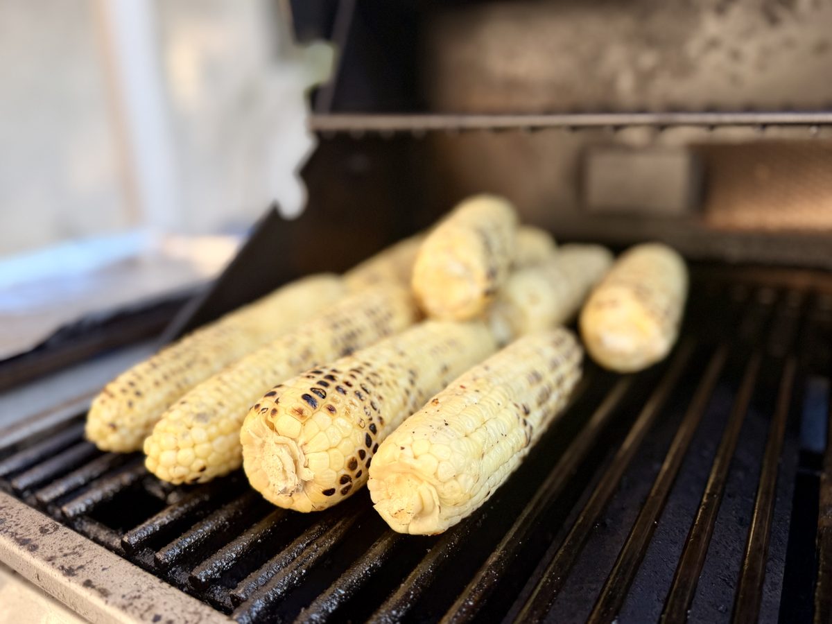 A close row of husked corn cobs with golden and blackened grill marks resting on the black grates of a grill, hood open behind.