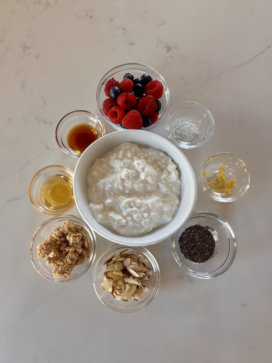 Overhead flat-lay on white marble of a white bowl of cottage cheese ringed by small glass bowls of raspberries and blueberries, ho