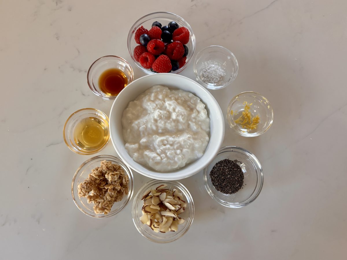 The same cottage cheese bowl surrounded by small glass bowls of berries, honey, vanilla, granola, almonds and chia seeds on white 