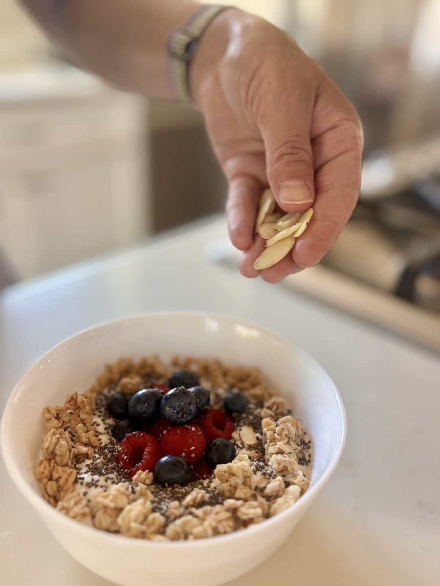 A hand sprinkling sliced almonds over a finished white bowl of cottage cheese ringed with granola and topped with blueberries, ras