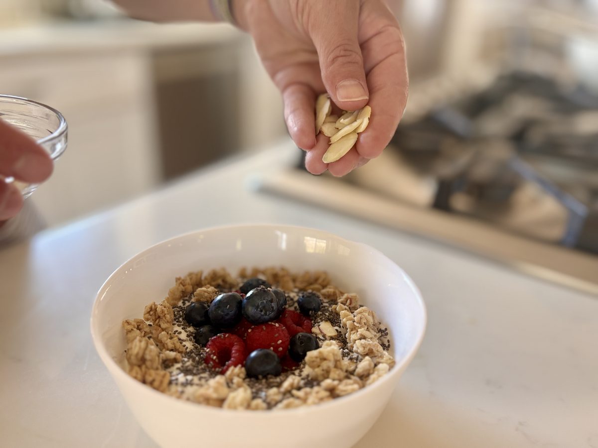 A white bowl of granola with cottage cheese, blueberries, raspberries and chia on a white counter, with a hand above it sprinkling