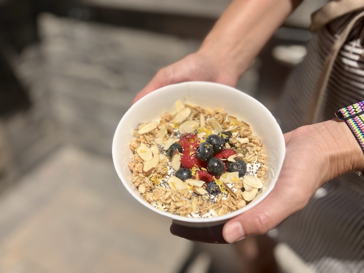 Hands holding out a finished white bowl of granola, cottage cheese, sliced almonds and mixed berries, blurred outdoor patio furnit