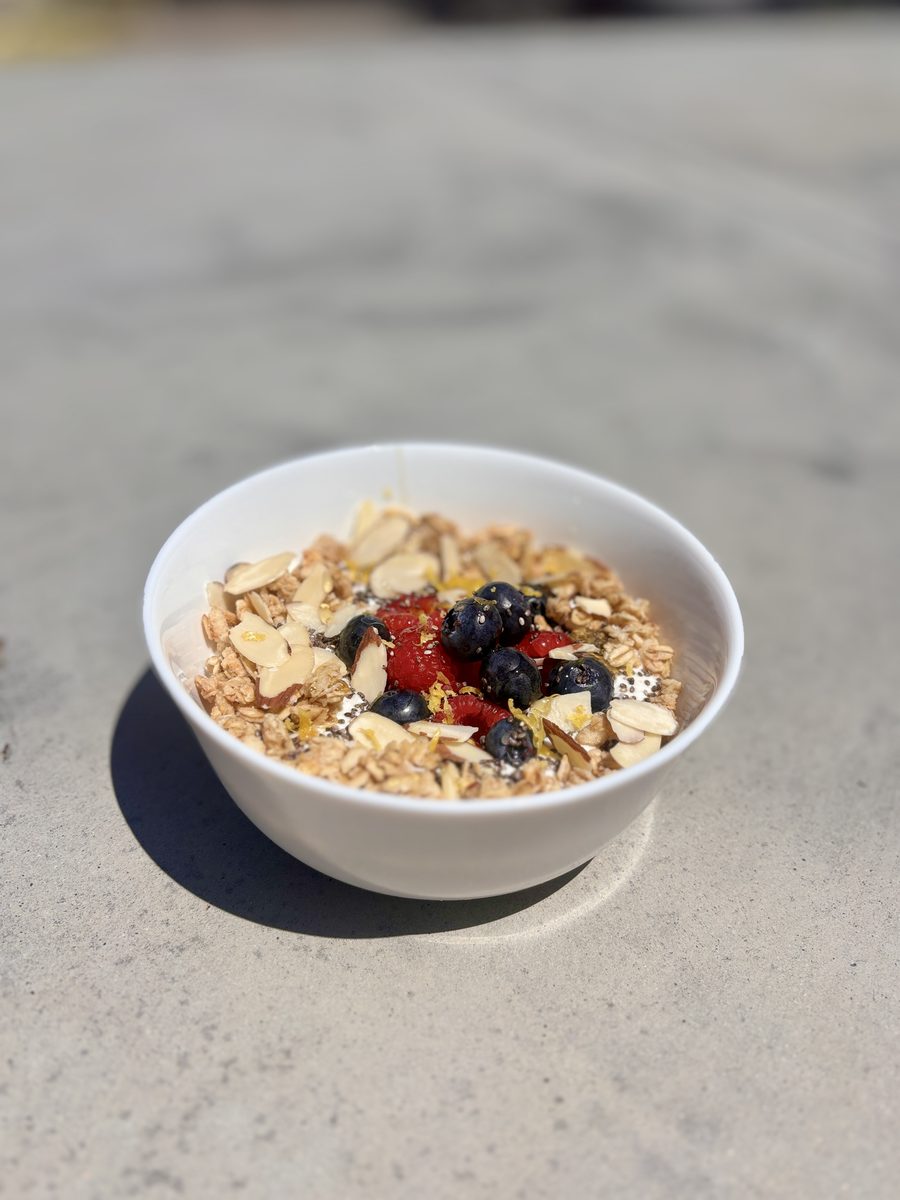 A finished white bowl of granola with cottage cheese, sliced almonds, strawberries and blueberries resting on a grey concrete outd
