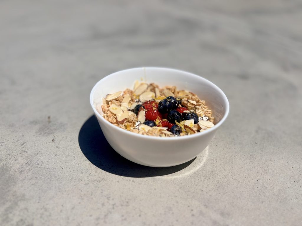 The same finished granola and berry bowl on a concrete surface in harsh direct sun, casting a hard shadow.