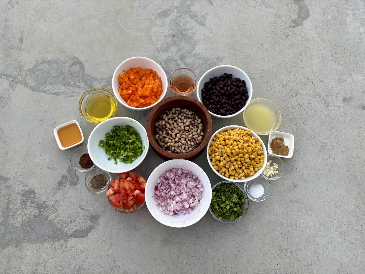 Overhead flat-lay on gray concrete of cowboy caviar components in white and glass bowls: diced orange bell pepper, black beans, bl