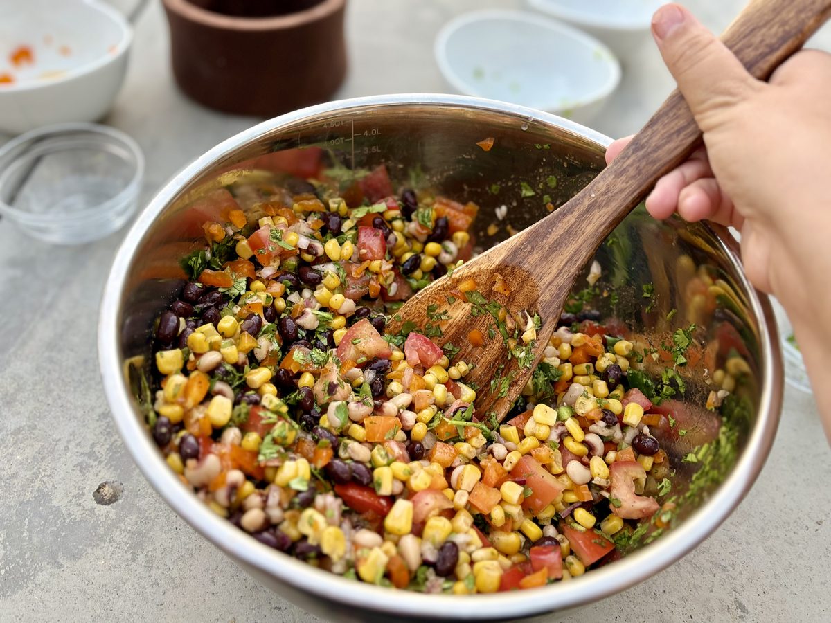 A hand stirring finished cowboy caviar — black beans, corn, black-eyed peas, diced tomato, red onion and cilantro in dressing — wi