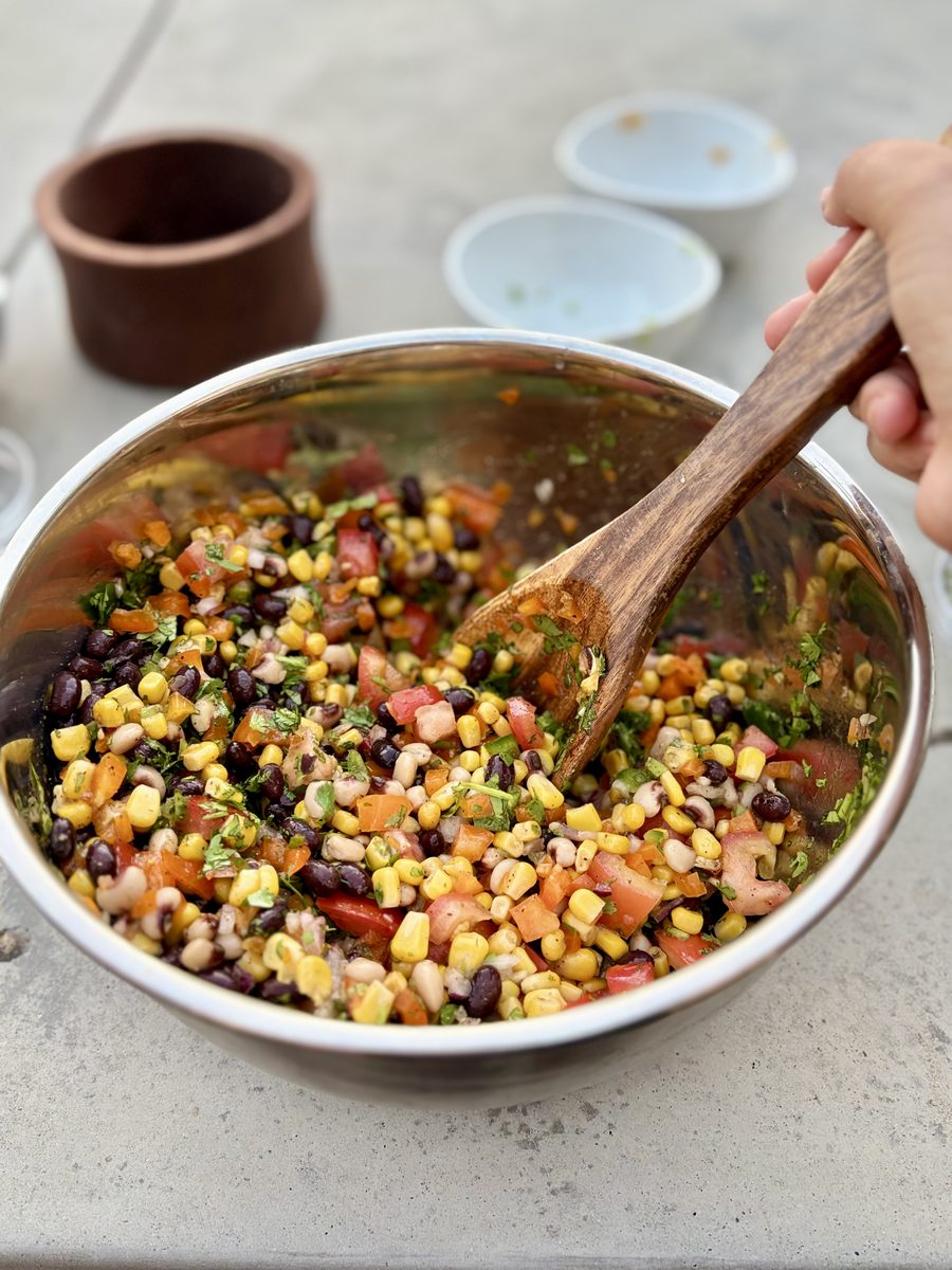 Overhead view of cowboy caviar being tossed with a wooden spoon in a stainless bowl, a hand on the spoon, small crocks and bowls b