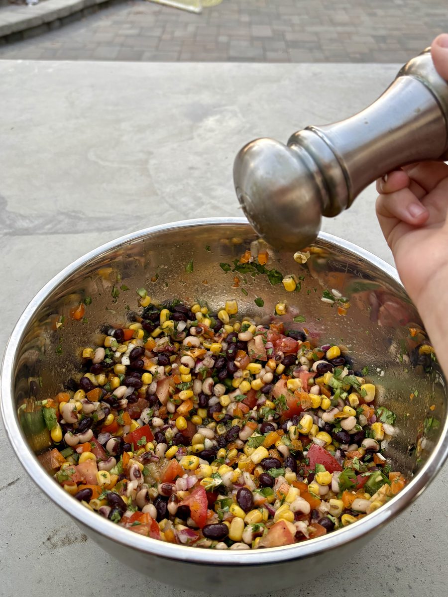 A hand grinding pepper from a metal pepper mill over a stainless bowl of mixed cowboy caviar on a concrete counter, shot from abov