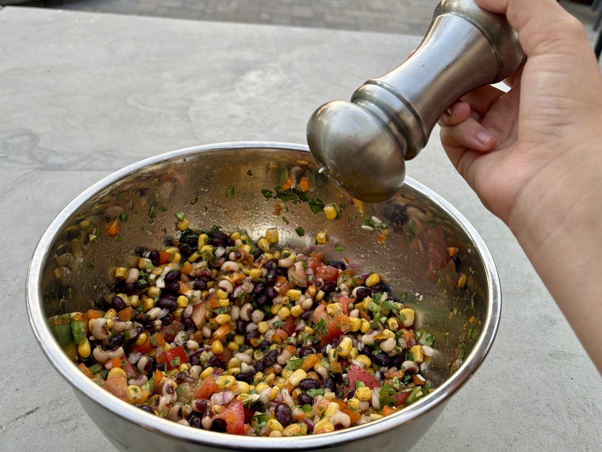 Close overhead view of a hand grinding pepper over a large stainless bowl of cowboy caviar — black beans, corn, tomato, onion and
