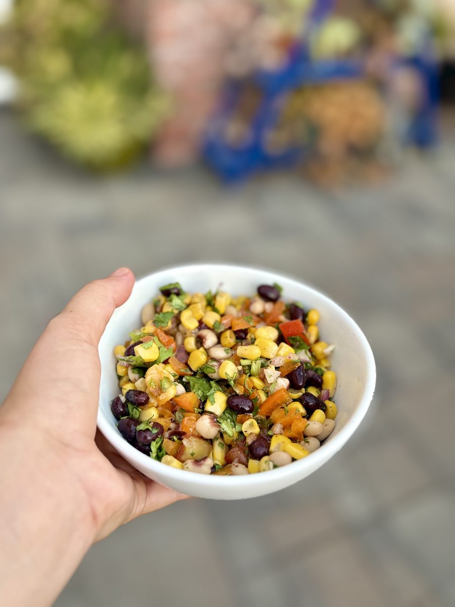 A hand holding a small white bowl of finished cowboy caviar — black beans, corn, tomato, pepper and cilantro — over a blurred pave
