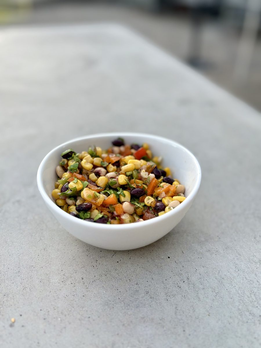 A small white bowl of finished cowboy caviar sitting on a smooth grey concrete outdoor table, patio blurred in the background.