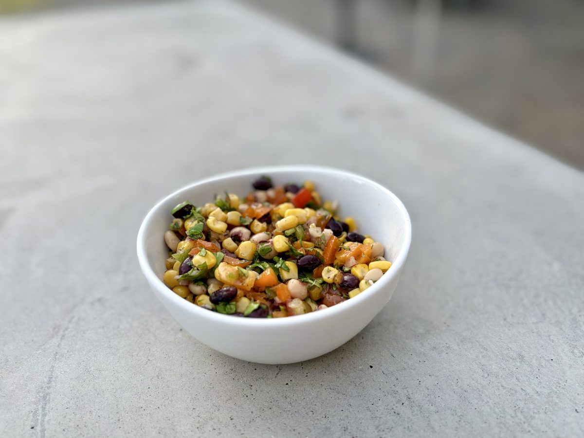 Closer shot of the finished cowboy caviar in a white bowl on a grey concrete counter, corn, black beans, tomato and cilantro clear