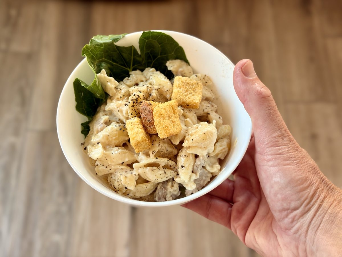A hand holding a white bowl of creamy cavatappi pasta with pieces of chicken, croutons, fresh spinach and black pepper, wood plank