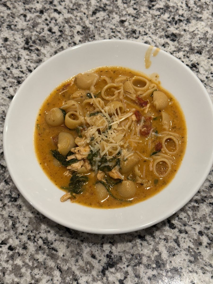 A white bowl of brothy soup with small pasta rings, white beans, greens and grated cheese on a granite counter; pale shreds in the