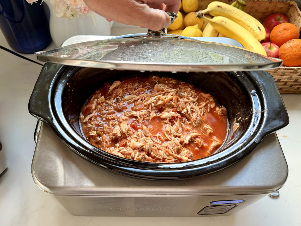 An open stainless slow cooker on a counter filled with shredded chicken in a red tomato sauce, a hand lifting the glass lid, with