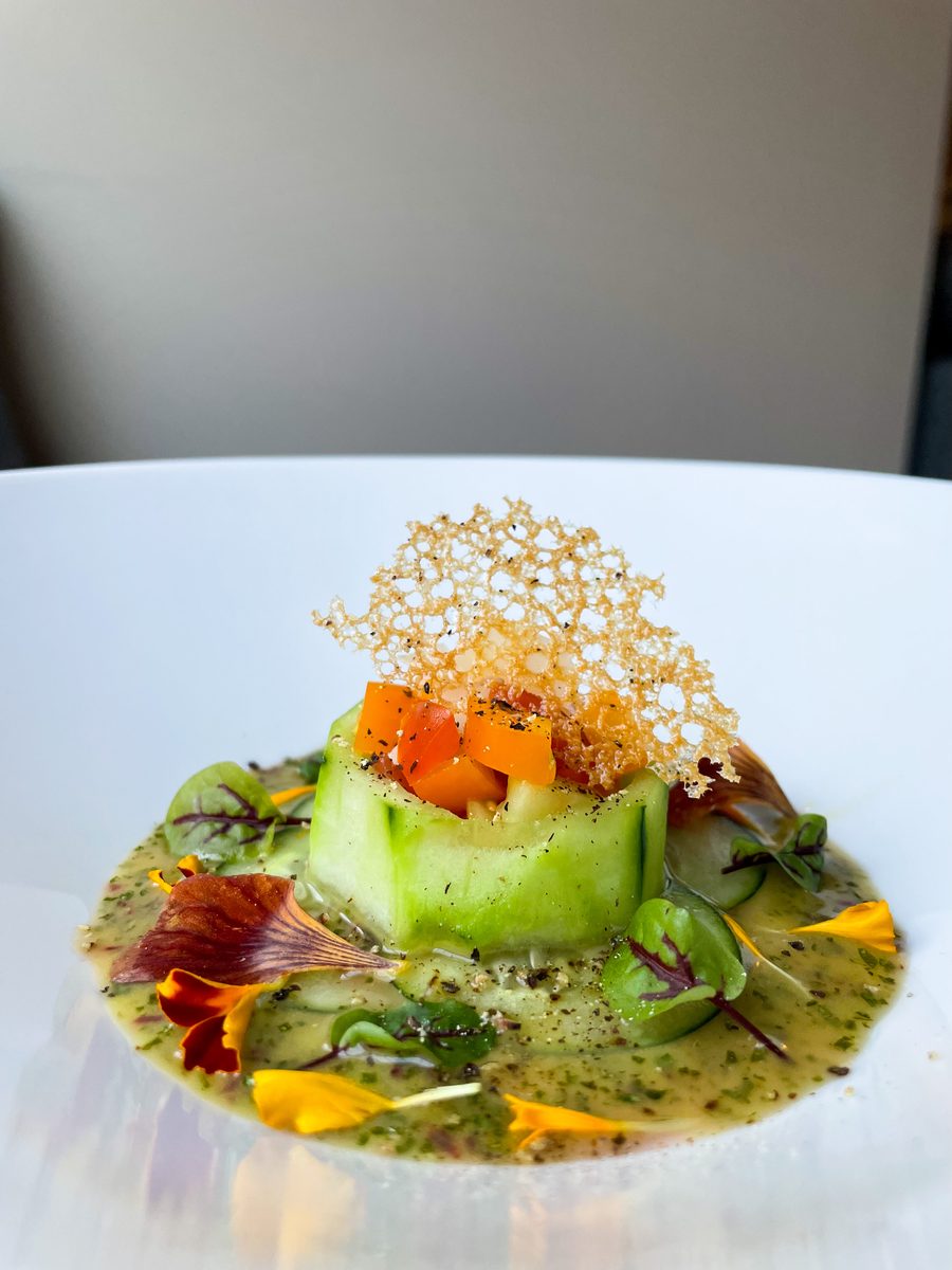 A fine-dining amuse-bouche on a white plate: a cucumber ribbon ring filled with diced melon and papaya, crowned with a lacy crisp 