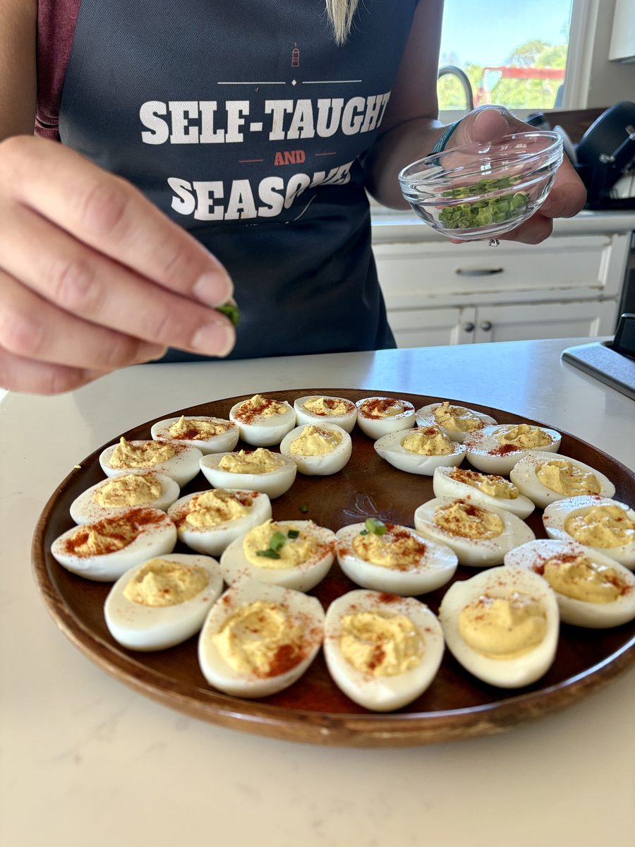 A round wooden platter of paprika-dusted deviled eggs on a kitchen counter while a hand sprinkles chopped herbs from a small glass