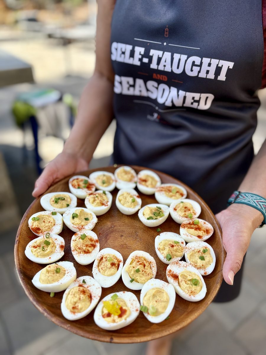 A person in a dark graphic apron holds a round wooden platter arranged with deviled eggs topped with paprika and chives, outdoor p