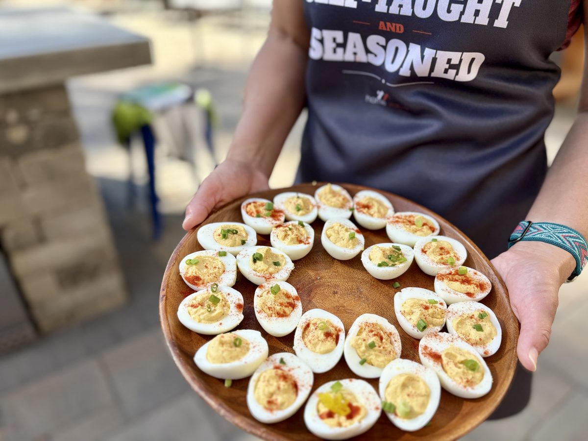 Two hands hold a wooden platter of deviled eggs toward the camera; the cook's lettered apron and an outdoor patio with stools fill