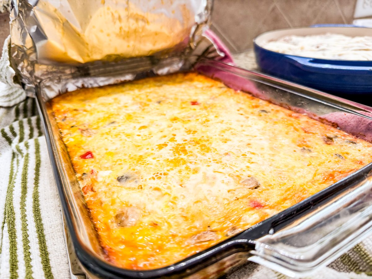 An angled close view of a cheesy baked egg casserole in a glass dish on a striped towel, cropped at the edges with a blue plate an