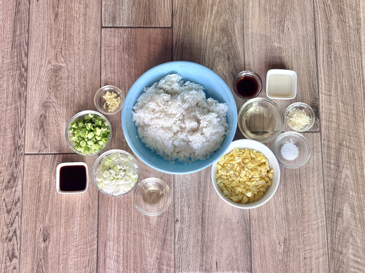 Overhead flat-lay on wood plank of egg fried rice components — a blue bowl of cooked white rice surrounded by small bowls of scram