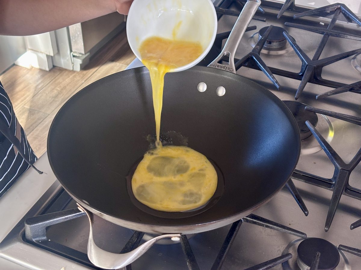 Beaten egg being poured from a white bowl into a hot black wok on a gas range, the egg already spreading into a round pool.