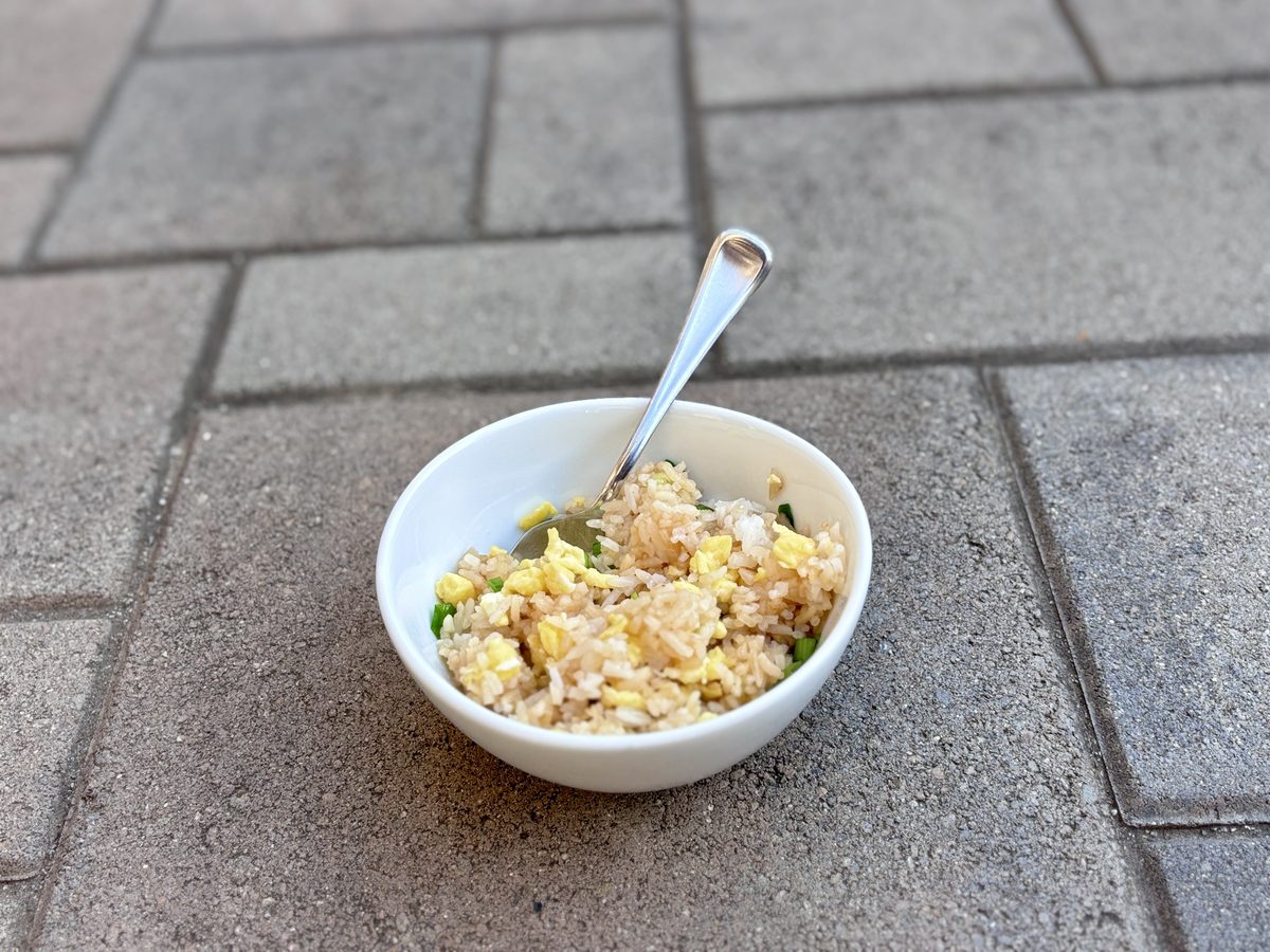 A finished white bowl of egg fried rice with a spoon standing in it, set on grey paving stones outdoors; yellow egg and green onio