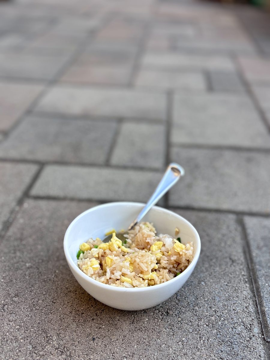 Pulled-back shot of the same served bowl of egg fried rice with a spoon sitting small in frame on outdoor pavers.