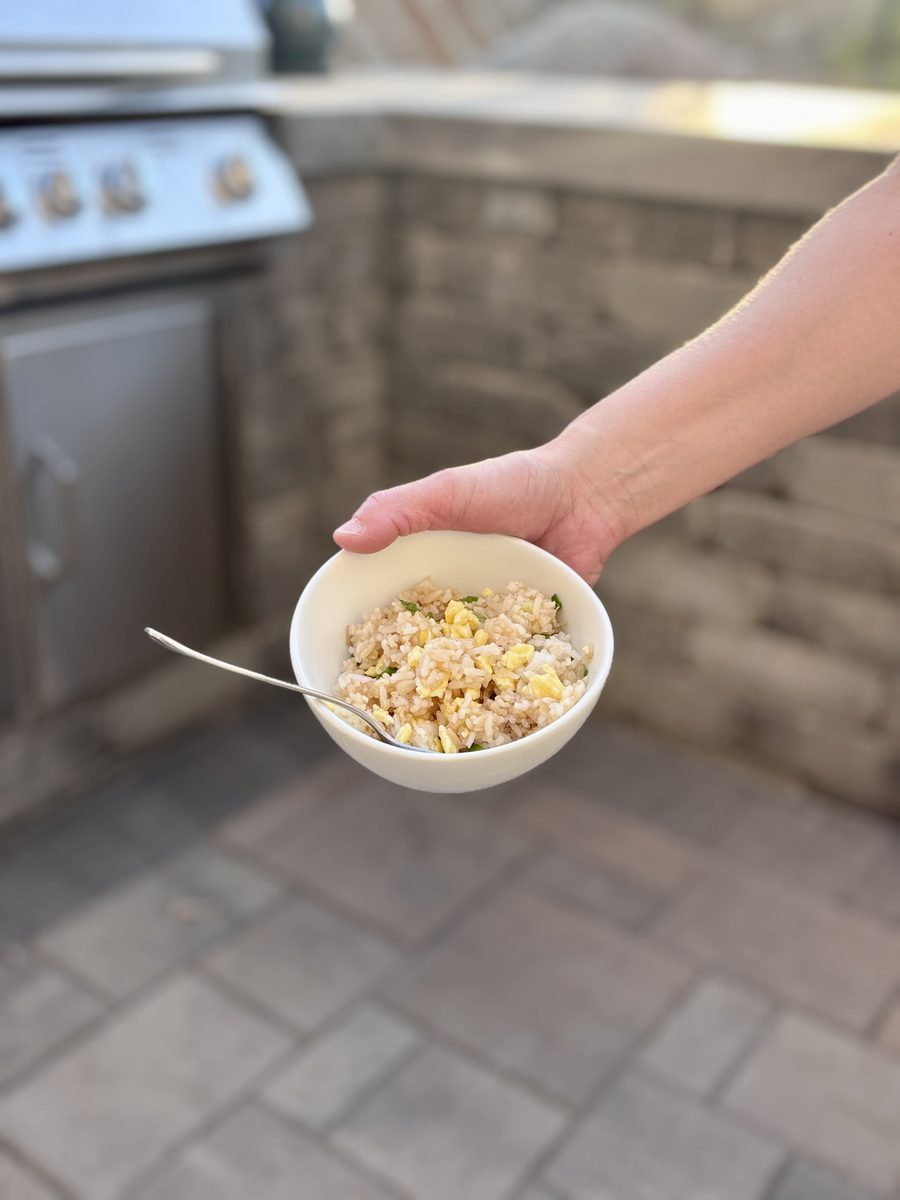 A hand holding a small white bowl of fried rice with a fork outdoors on a patio; no meat is visible in the rice.