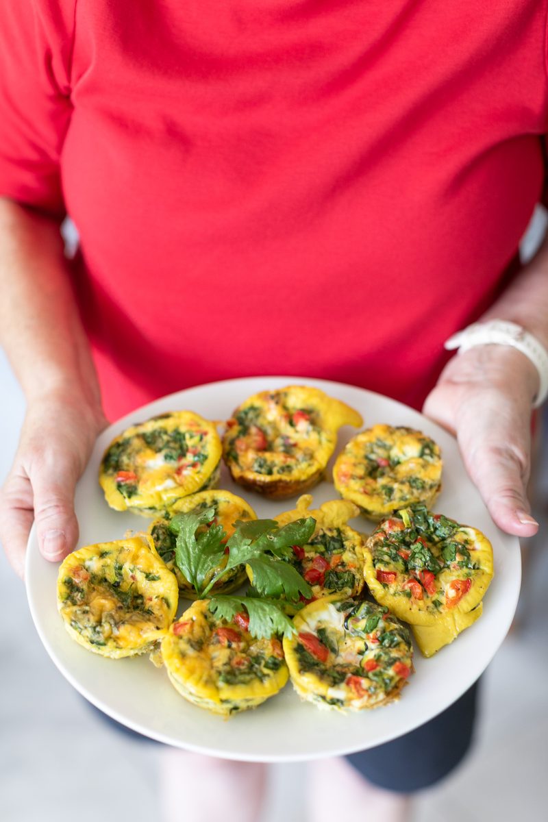 A woman in a red shirt holds a white plate of baked egg muffins garnished with a sprig of cilantro, outdoors in bright light.