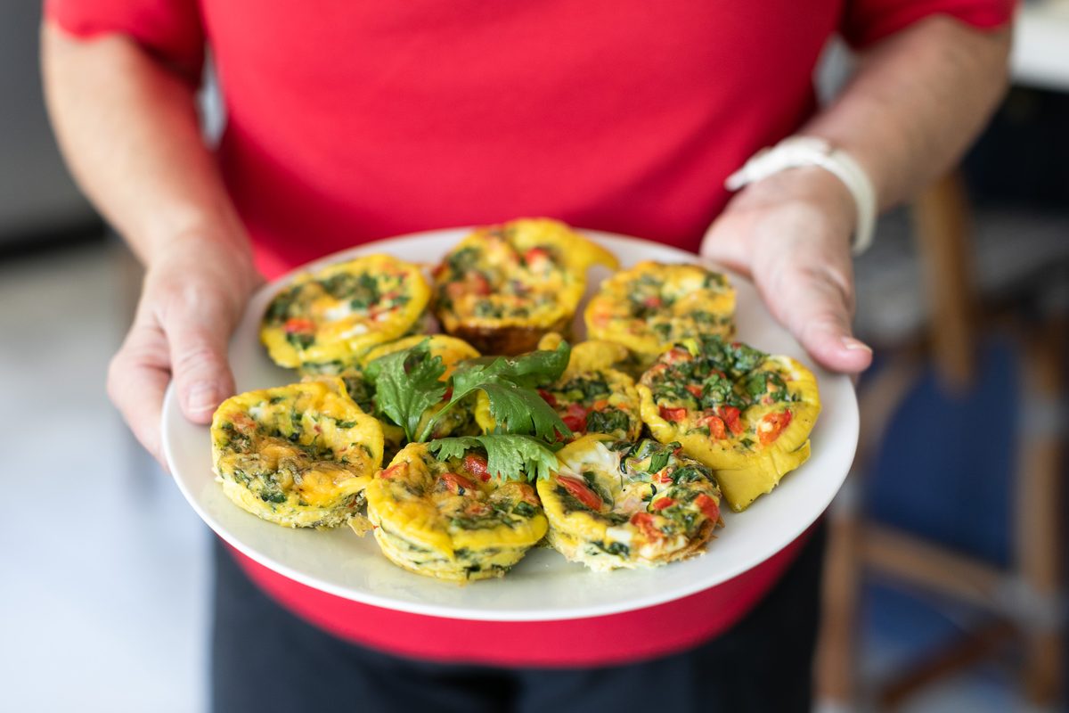 A person in a red shirt holds out a white plate of baked egg muffins topped with a cilantro sprig, blurred kitchen background.