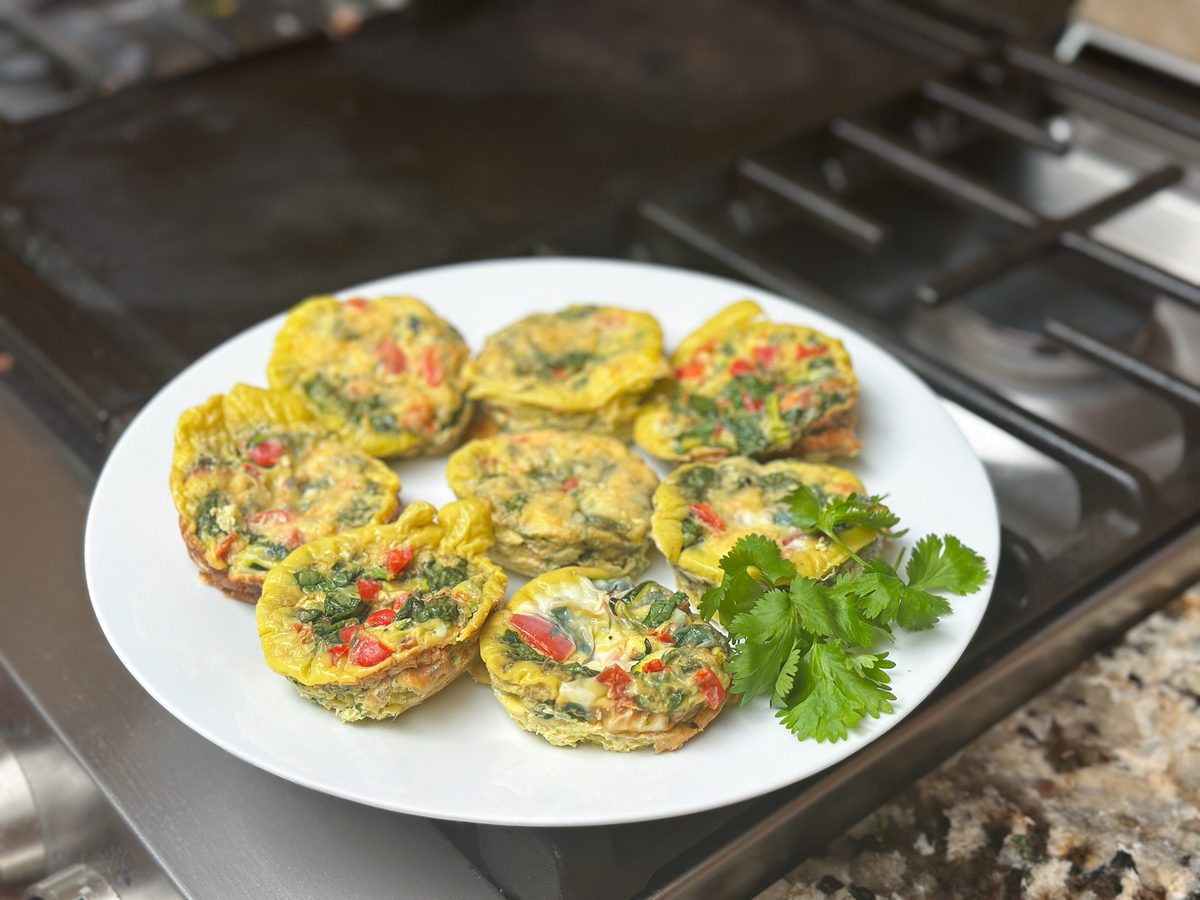 A white plate of baked egg muffins with spinach and red pepper, garnished with cilantro, resting on a counter beside a gas cooktop