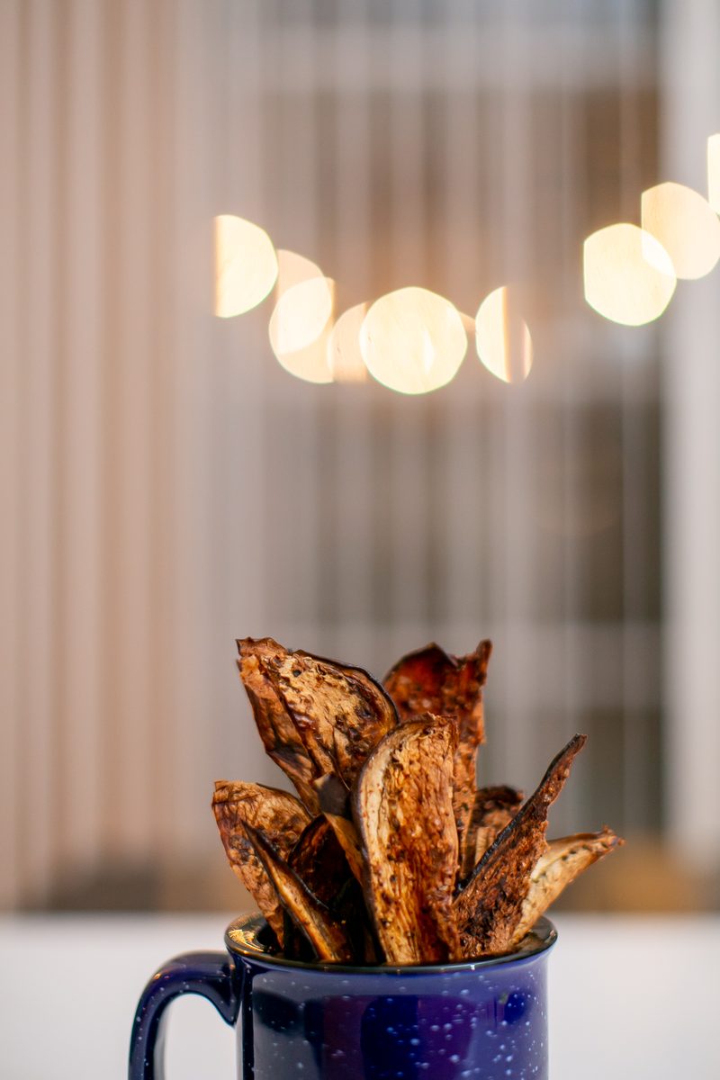 Crisp baked eggplant chips standing upright in a dark blue enamel mug on a white table with warm bokeh lights behind.