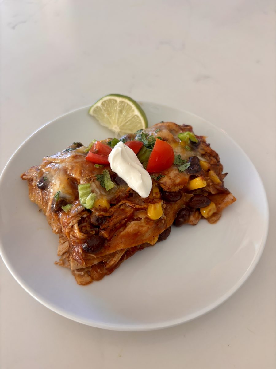 A square slice of layered enchilada casserole with corn, black beans and melted cheese on a white plate, topped with sour cream, d