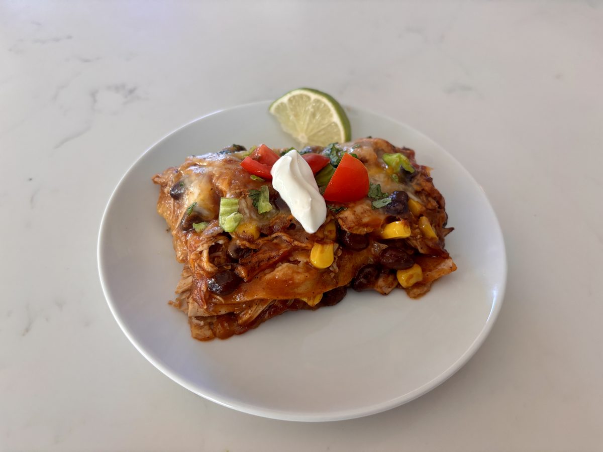 A slice of layered enchilada casserole with corn, beans, cheese and red sauce on a white plate, garnished with sour cream, tomato,