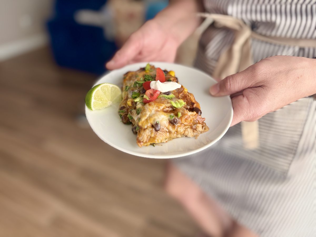 Hands hold a small white plate with a slice of layered tortilla casserole containing black beans, corn and cheese, topped with sou