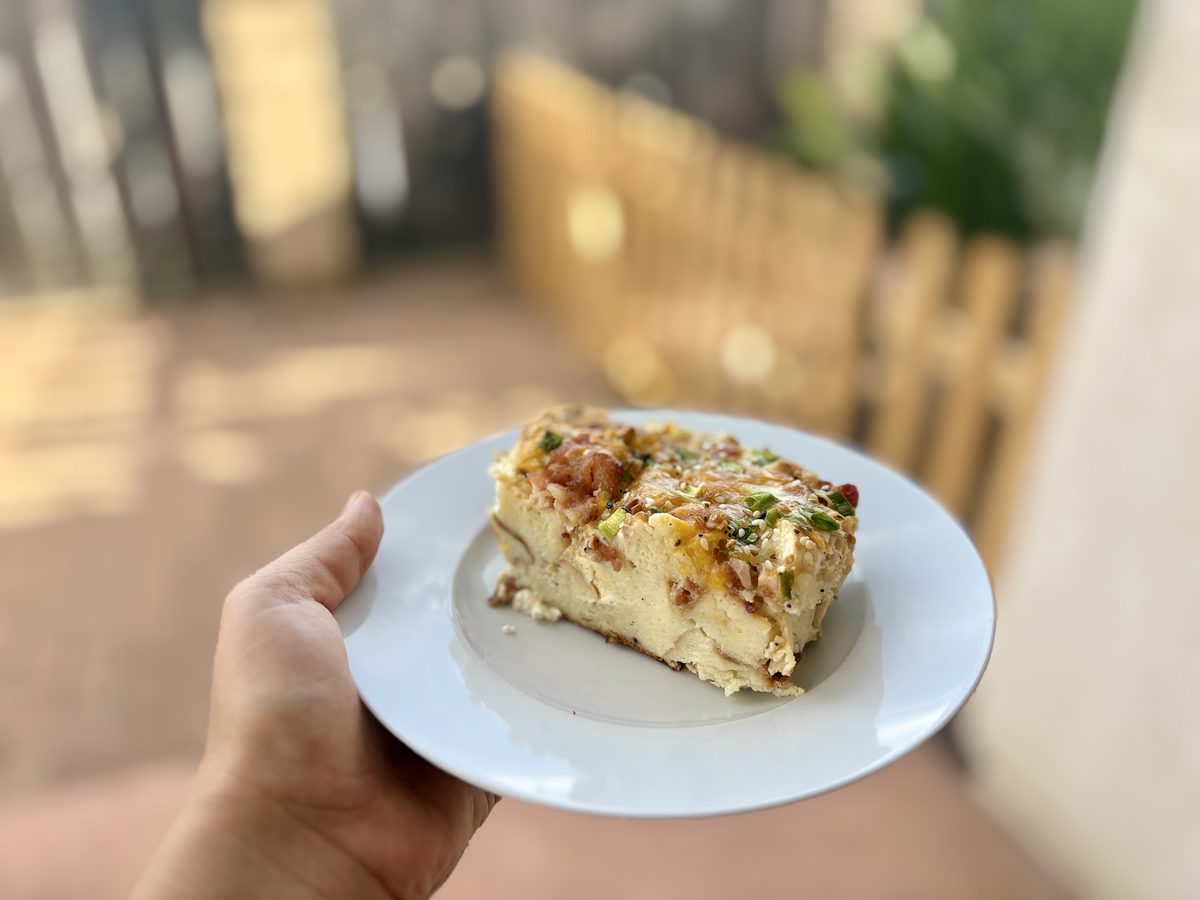 A hand holding a white plate with a square slice of layered egg-and-bread casserole with a browned cheesy top; the brown flecks in