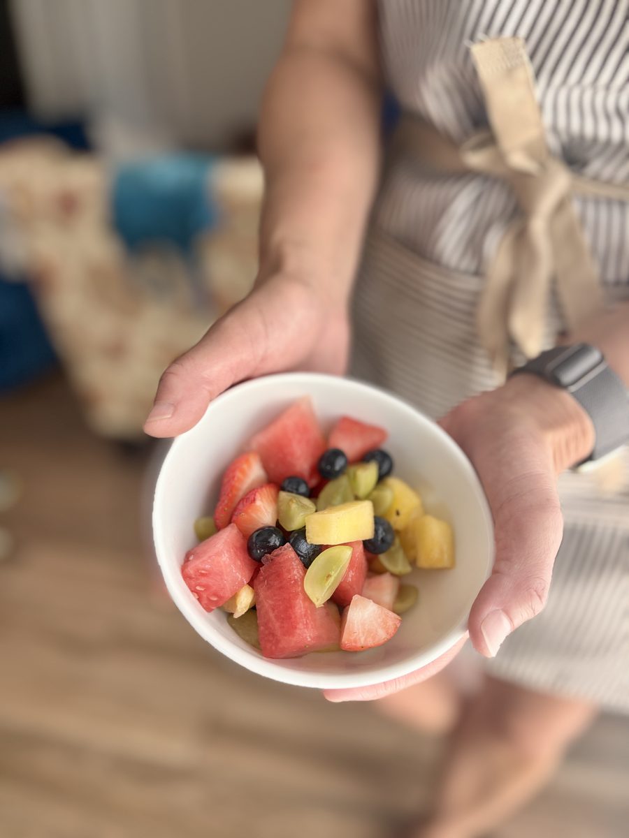 A hand holds a small white bowl of fruit salad with watermelon, pineapple, grapes and blueberries; person in a striped apron and a