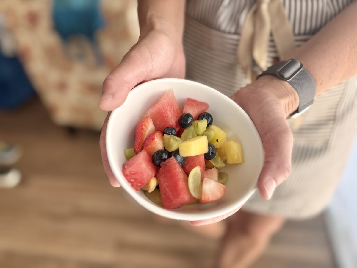 Two hands cup a white bowl of chunky fruit salad (watermelon, pineapple, grapes, blueberries) at chest height, person in a striped