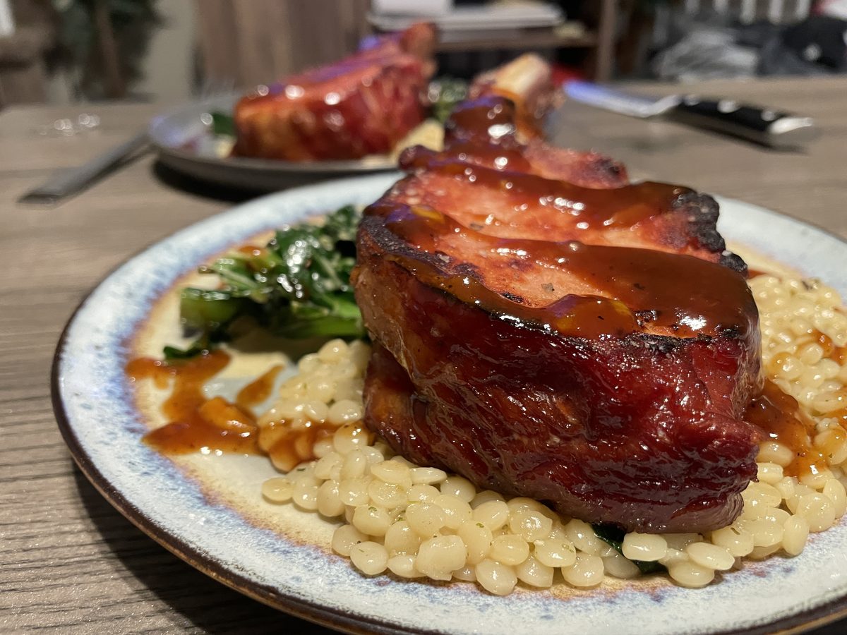 A thick glazed bone-in pork chop with dark sauce resting on a bed of small pasta or beans with greens behind it, on a speckled cer