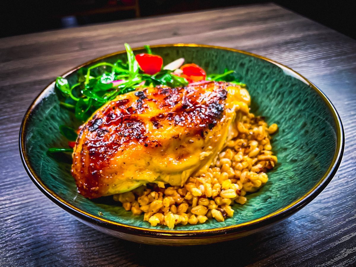 A caramelized glazed chicken breast on a bed of farro and corn with a tangle of greens and radish garnish, plated on a teal cerami