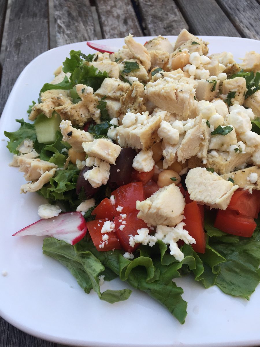 Chopped grilled chicken and crumbled feta piled over lettuce, tomato, cucumber, radish and olives on a white square plate on a wea