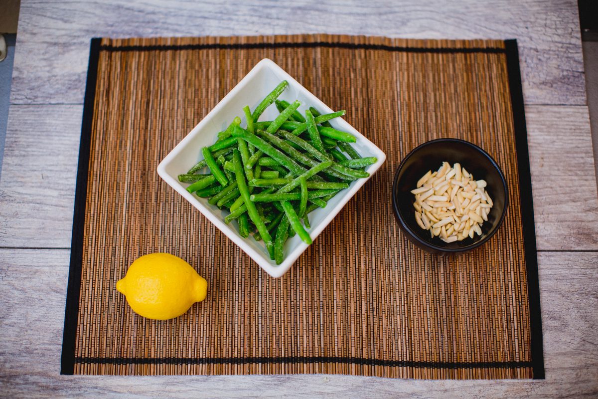 Overhead shot of green beans in a white square dish with a lemon and a small black bowl of slivered almonds on a bamboo placemat o